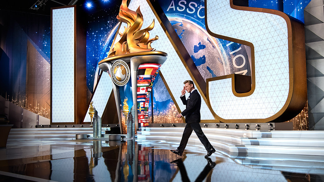 David Miscavige walking onstage at the International Association of Scientologists Event, in front of a backdrop featuring the IAS emblem and global flags