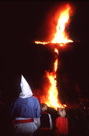 A cross on fire with a man in a white KKK cap watching